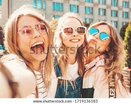 Three young smiling hipster women in summer clothes.Girls taking selfie self portrait photos on smartphone.Models posing in the street.Female showing positive face emotions in sunglasses