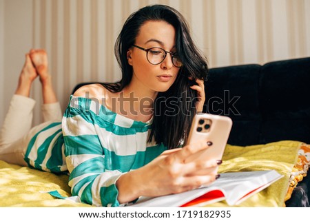 Female addicted from gadgets distracted from lesson at home, making self portrait. Young woman with beautiful smile taking selfie while reading book on the bed. 