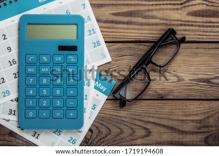 Calculator with sheets of the monthly calendar, glasses on wooden background. Economic calculation, costing. Top view Royalty-Free Stock Photo #1719194608