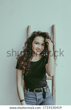A smiling european woman in a black t-shirt and blue jeans standing in front of the white wall and stairs on the background. A happy potter female. Ceramic studio concept
