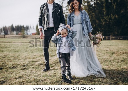 Stylish family in the autumn mountains. Stock photo. A guy in a leather jacket and a young girl in a gray-blue dress with his son on the background of the forest and wooden fence
