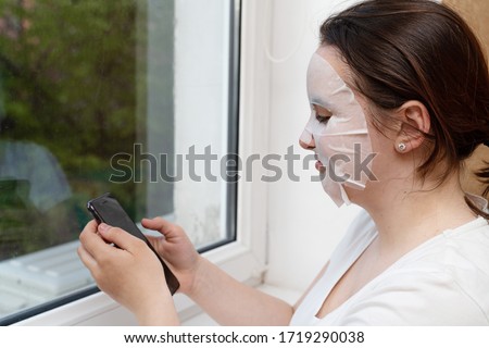 A young woman in a mask of cosmetic fabric is standing near the window in a white T-shirt and is surfing in a smartphone. The concept of quarantine, home care yourself