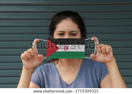 A woman with Sahrawi arab flag on hygienic mask in her hand and lifted up the front face on dark green background. Tiny Particle or virus corona or Covid 19 protection. Concept of Combating illness.