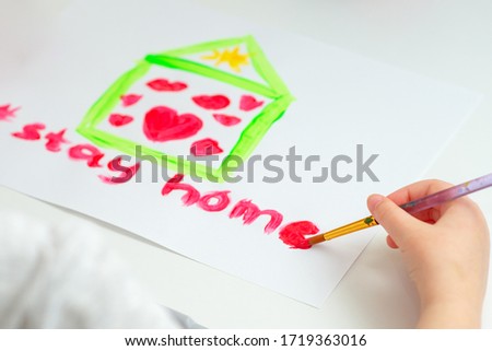 Child is writing words Stay Home with red watercolor under drawn house with hearts on sheet of paper. Stay Home concept.
