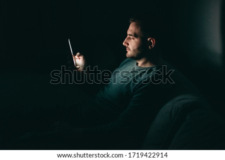 Young white man reads news on his phone at home at quarantine at night. Coronavirus, isolation, depression, crisis.