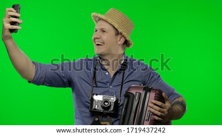 Portrait of man tourist with suitcase in hand is making selfies using mobile phone. Smartphone communication. Handsome man in blue shirt and hat with retro camera. Chroma key. Green screen