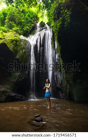Young Caucasian woman standing near the waterfall, meditating, practicing yoga and pranayama. Hands in namaste mudra. Concentration and relaxation. Krisik waterfall in Bangli, Bali, Indonesia.
