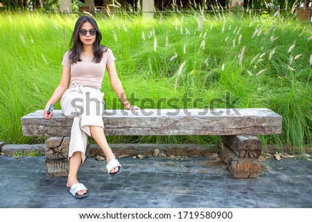 Beautiful Asian woman is sitting on a wooden chair with  green grass background. Portrait of asian girl is looking at camera.
