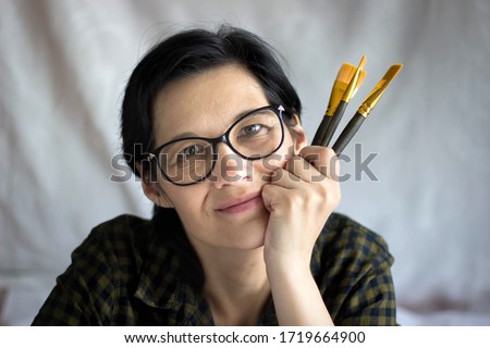 A beautiful smiling brunette woman with glasses is an artist with brushes in her hand, happily looking at the camera, on a light background. The concept of creativity.