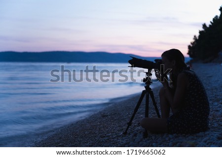 Young female photographer sitting on pebble beach in the evening with her camera set on tripod ready  to shoot the evening sea and landscape.