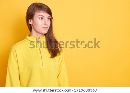 Image of charming woman with perfect skin and pleasant appearance, posing isolated over yellow studio background and looking aside, lady wearing hoodie. Copy space for advertisement or promotion.