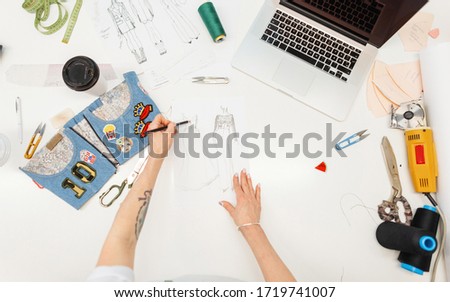 Fashion designer working in studio. Close up design. Work laptop, coffee, thread, electric cutter, sketches, denim, centimeter and other tools for sewing on the desktop, top view