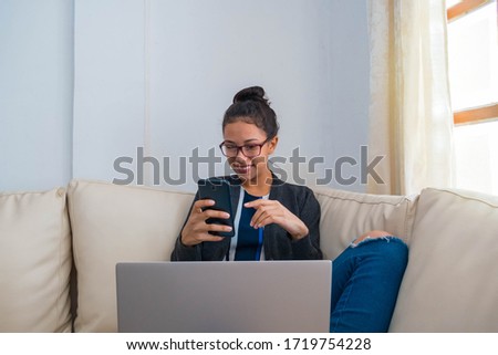 A young woman makes purchases on the Internet. The freelancer pays using the smartphone. The happy girl enjoys shopping and sitting by the window with a laptop at home
