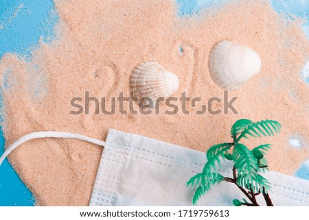 travel and quarantine concept, inscription on sand 2020, plastic toy miniature palm, protective medical face and shell mask, blue background, coronavirus pandemic