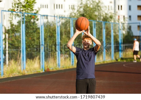 Cute boy in blue t shirt plays basketball on city playground. Active teen enjoying outdoor game with orange ball. Hobby, active lifestyle, sport for kids.
