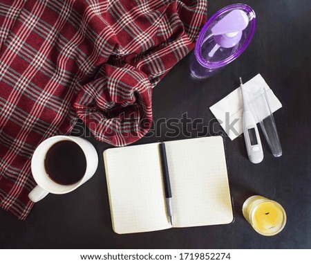 Black table with a checkered jacket, cup of coffee, blank notebook with a pen, hand sanitizer, and a temperature measuring device. Concept for work from home during the corona virus pandemic. 
