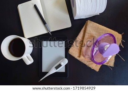 Black table with a checkered jacket, a cup of coffee, a blank notebook with a pen, hand sanitiser, tissue paper, and a temperature measuring device. The concept for work from home during the pandemic.
