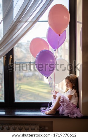 Little girl at home sitting by the window and holding balloons in his hand. Image with selective focus.