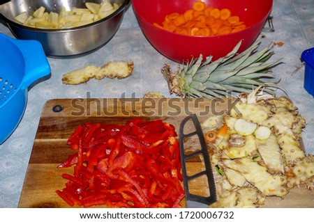 Artisanal preparation of a homemade mixed vegetable dish in a kitchen : fresh red peppers cut into slices, peelings of pineapple and a knife with hollow ground blade on a wooden chopping board Royalty-Free Stock Photo #1720006732