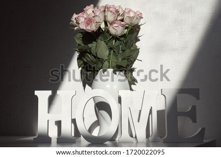 Wooden and decorative letters of the word home in front of a vase of roses. Sunlight and shadows on the wall behind. Home is where your heart is.