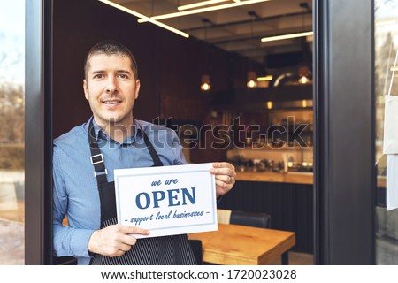 Small business owner smiling while holding open sign of restaurant reopening after lockdown quarantine due to coronavirus pandemic - Entrepeneur opening cafeteria activity to support local businesses. Royalty-Free Stock Photo #1720023028