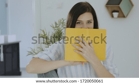 Portrait of happy young brunette woman smiling and holding  yellow daily planner at home. Caucasian girl looking at camera and covering his face with a book.
