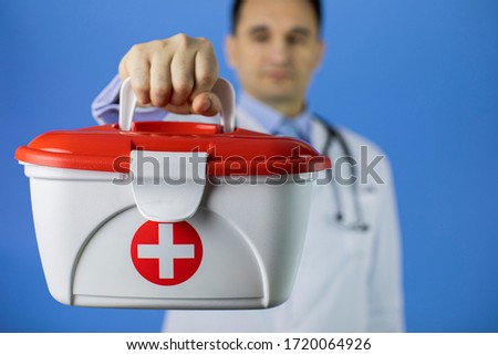handsome dark hair 40s male doctor holds red cross medical aid kit selective focus isolated on blue background copy space. emergency concept