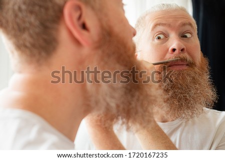 Attractive middle-aged man cutting his moustache and beard by himself in front of mirror at home. Beard care during quarantine. Coronavirus outbreak, lockdown, staying home on self-isolation. Closeup