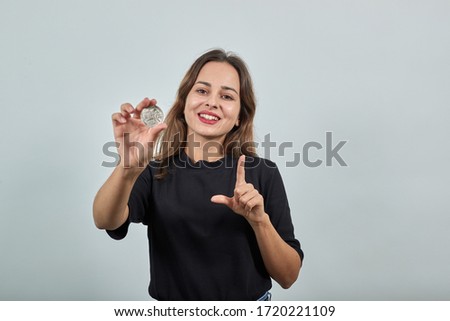 Cute Young Woman Brunette In Black T-Shirt, Blue Jeans With Belt On Gray Background, Happy Girl Smiling Holds A Silver Bitcoin Coin And Gestures With Her Fingers