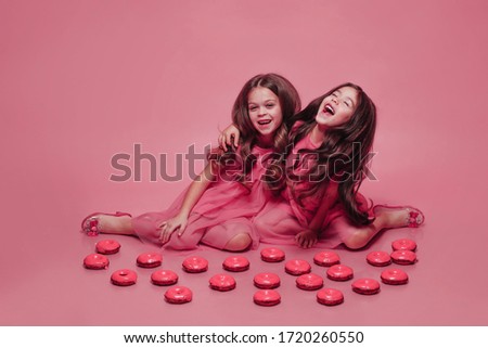 Girls in pink dresses with long hair, sitting on the floor among a lot of doughnuts. The model's friends laugh and hug.