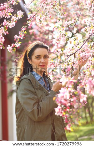 A young woman in a green jacket stands and rejoices near a blossoming pink tree on a sunny day.
The picture was taken with selective focus. Noise effect on the photo.