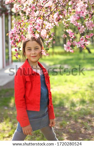 A young girl in a red jacket stands near a blossoming pink tree on a sunny day.
The picture was taken with selective focus. Noise effect on the photo.