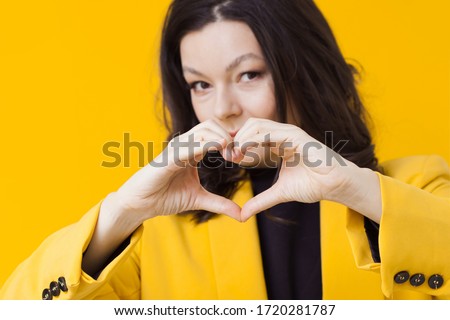 A young brunette woman in a yellow jacket shows a heart gesture. A sign of love and support. Positive portrait on a yellow background