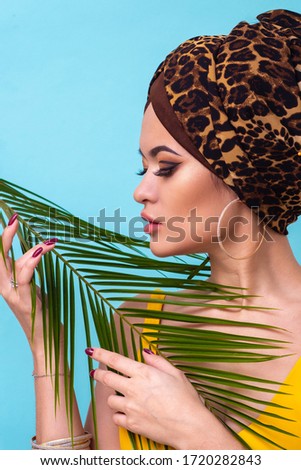 An attractive young woman in a stylish turban made of leopard print fabric on a blue background. Girl with bright makeup. Beach style, palm branch and heat. Vertical format stories