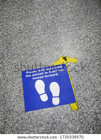 'Please wait here until the person in front has moved forward' footprint sign on the floor of a local grocery store during the COVID pandemic in the UK illustrating concept of social distancing