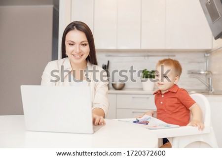 Woman on maternity leave working home online with laptop in kitchen with small child. Concept mom work while in quarantine isolation during Covid-19.