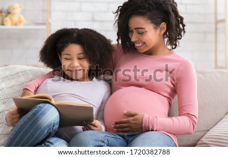 Cute Little Black Girl Reading Book With Her Beautiful Pregnant Mom At Home, Embracing On Couch And Smiling, Closeup