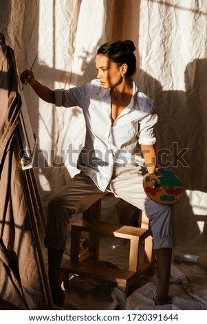 At sunset, a beautiful girl paints a picture with a palette in her hands, in a bright studio, in a white shirt and beige pants