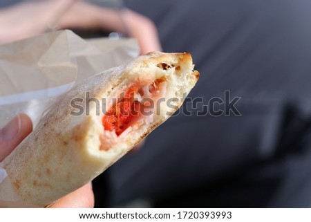 Eating ham & mozzarella calzone in a park. This calzone pizza from a grocery store in Switzerland. Person is holding the calzone in his hand. Closeup of the filling - tomato sauce, cheese & meat.