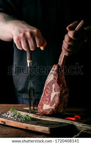 The chef's hands hold a raw Tomahawk a beef steak and a meat fork over a chopping Board