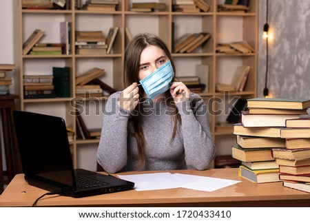 a young girl sits at work in an office and takes off a medical mask. quarantine end. bookcase on the background. Piles of books and a laptop are nearby. Royalty-Free Stock Photo #1720433410