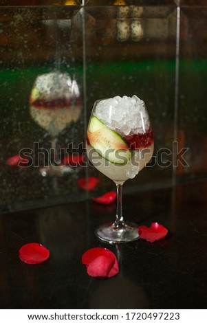 close up drink shot of a delicious fresh yellow red cocktail with ice, a cucumber slice in a transparent wine glass standing on a table with red rose petals, reflected in a dirty mirror in the back