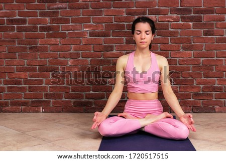 Beautiful young yogi woman in pink clothes practicing yoga in lotus position. Girl meditating with eyes closed against the brick wall background for copy space