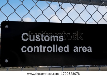 A black metal display sign with customs controlled area written in white letters. The sign is attached to a metal chain link wire fence. The background has blue skies and white clouds. 