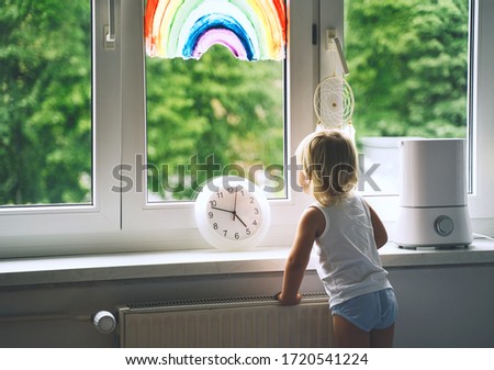 Little girl looks out window at spring time. Cute child on background of painting rainbow on window. Kids leisure at home, safety joy symbol, time concept. Support during quarantine