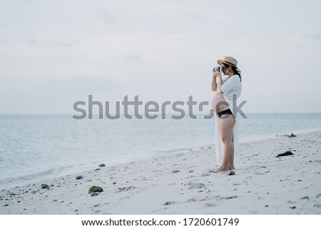Beautiful asian young pregnant woman with white dress is standing to take a picture of the sunset by the sea at Phangan island in evening time. Blur sea background.split color.