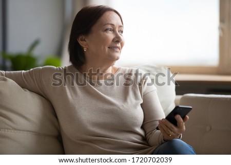 Dreamy middle aged woman resting on couch, holding phone, waiting for call or distracted from chatting online in social network, smiling older female looking in distance, enjoying leisure time Royalty-Free Stock Photo #1720702039