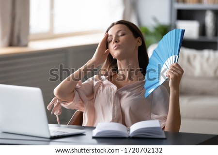 Overheated woman waving fan, sitting at desk with laptop at home, stressed young female suffering from heating at home, feeling discomfort, hot summer weather or fever, heatstroke concept Royalty-Free Stock Photo #1720702132
