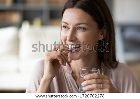 Close up smiling woman taking white round pill, holding water glass in hand, happy young female taking supplement, daily vitamins for hair and skin, natural beauty, healthy lifestyle