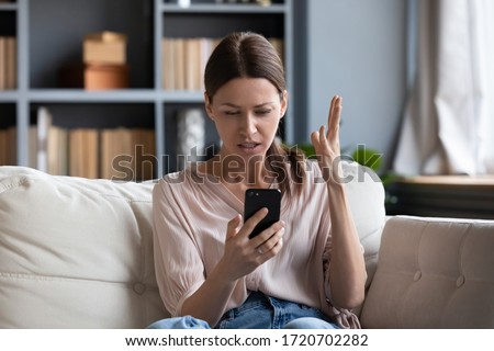 Confused angry woman having problem with phone, sitting on couch at home, unhappy young female looking at screen, dissatisfied by discharged or broken smartphone, reading bad news in message Royalty-Free Stock Photo #1720702282